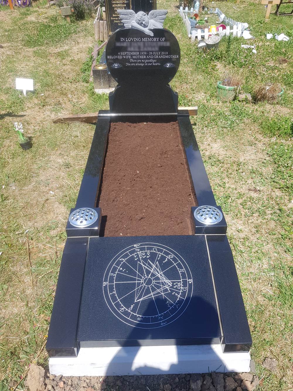 Black Granite heart shaped headstone with hand carved angel overlooking upon kerb set with foot panel and two square vases, soil centre.